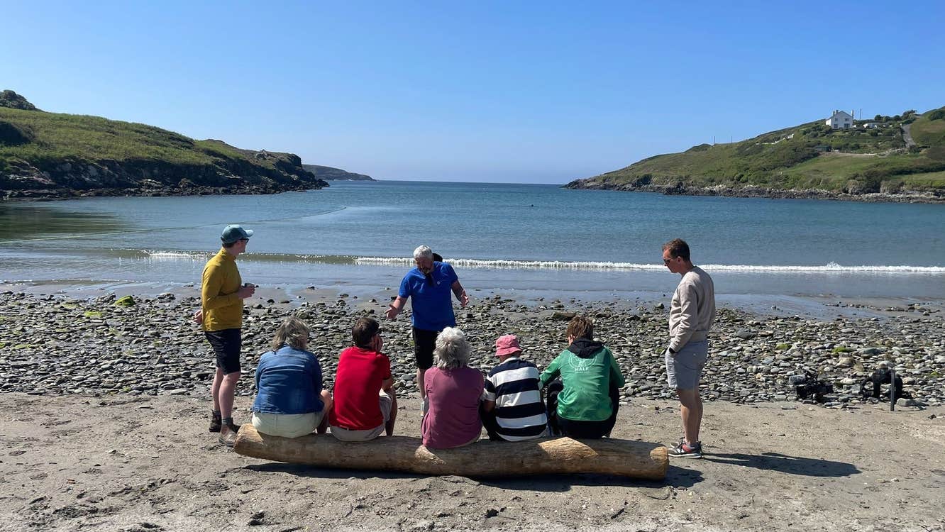 A group of people on a hiking tour sit on a log on a beach listening to their tour guide