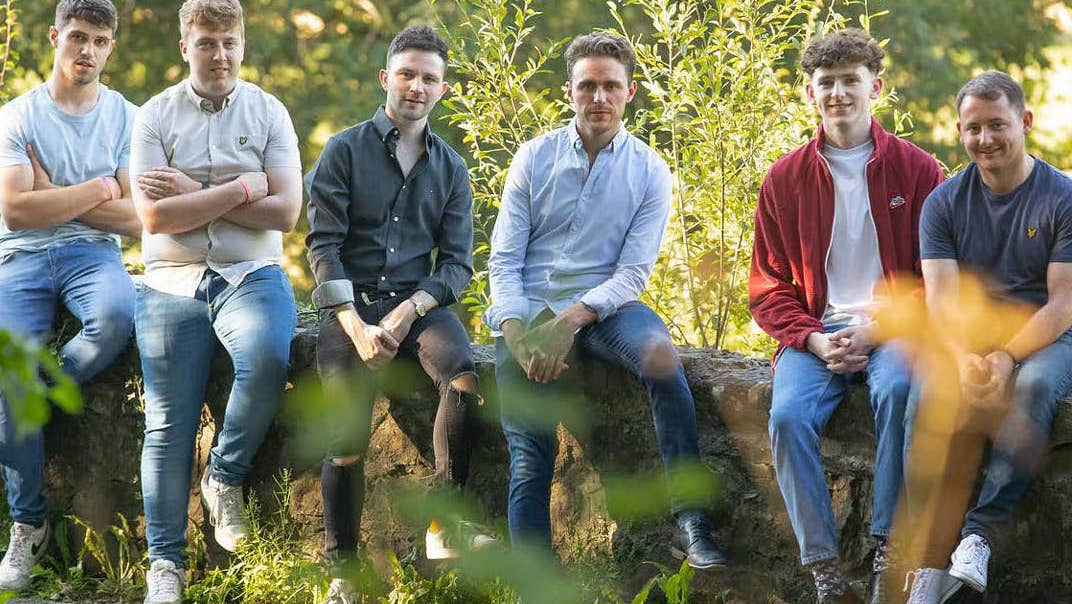 6 casually dressed men seated on a low stone wall surrounded by greenery