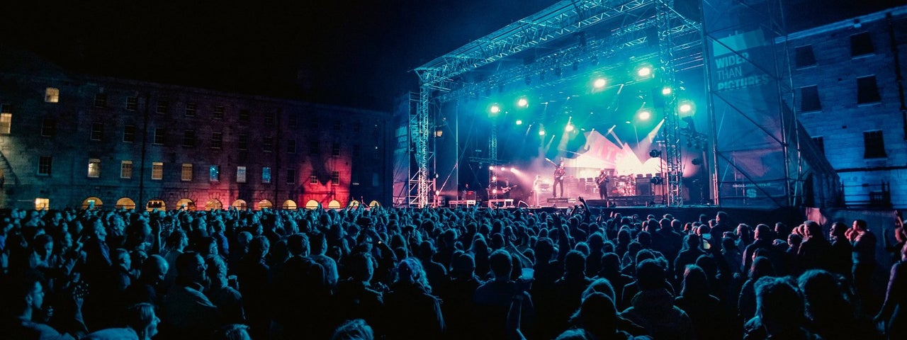 Distant view of large outdoor stage at night with large audience watching musicians