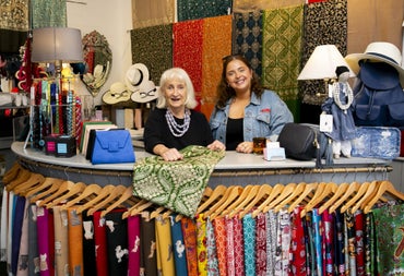 Two people behind a shop counter with scarfs on hangers hanging from a rail on it