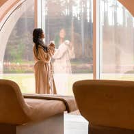 A lady in a bathrobe staring out a large window in a room with treatment beds
