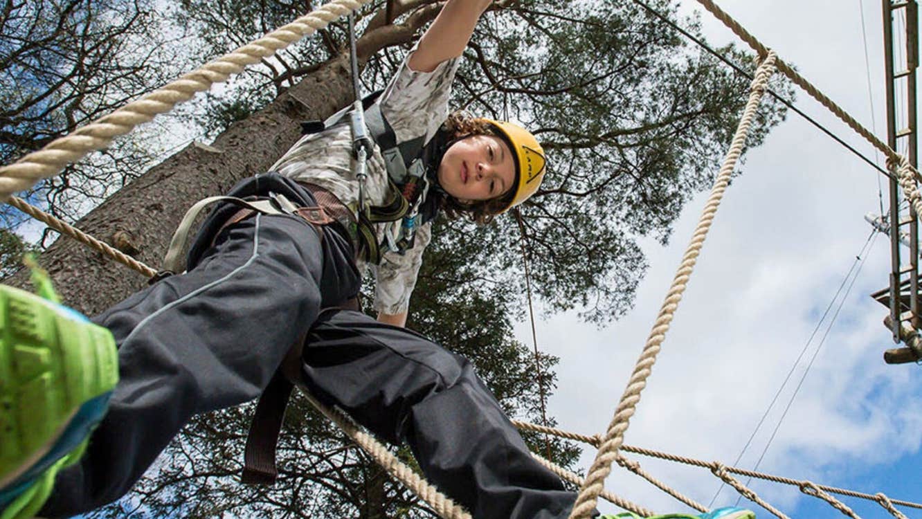 Child looking down at the camera walking on a rope between trees with a blue sky above
