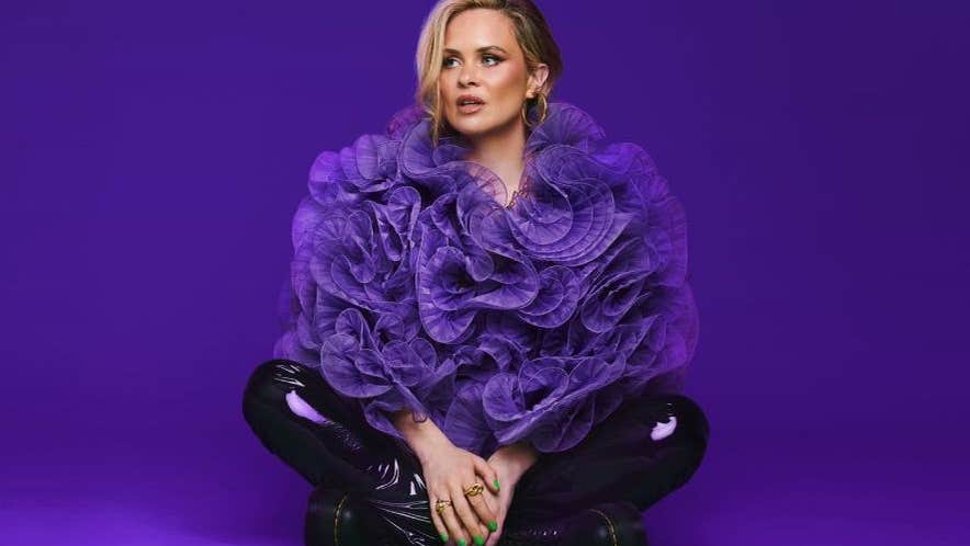 A woman is seated crossed legged on a purple floor wearing a huge, frilly purple top and dark shiny trousers.