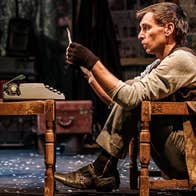A man seated in a chair frame looking at a piece of paper with old small typewriter on an old chair in front of him.