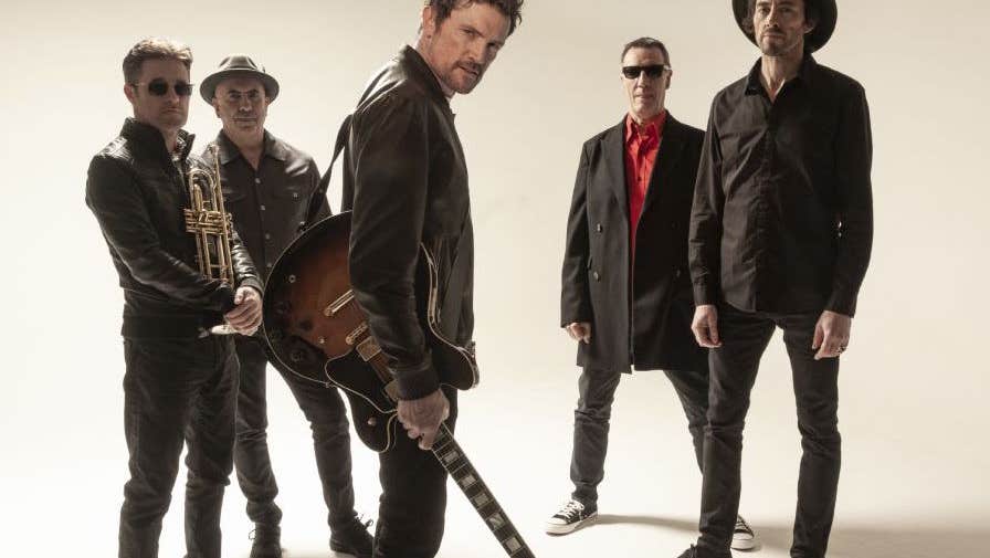 5 men dressed in dark clothes and leather jackets are standing in a casual group looking serious with one in the centre towards the front with a guitar swinging down looking questioningly
