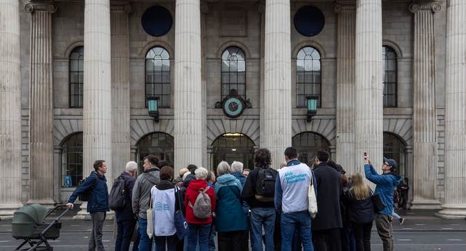 A group of people standing in front of the facade with pillars of grand building.