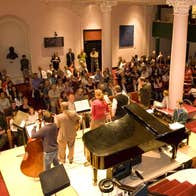 Viewed from behind and above, rear view of small stage with musicians playing and seated audience in a hallway with stairs
