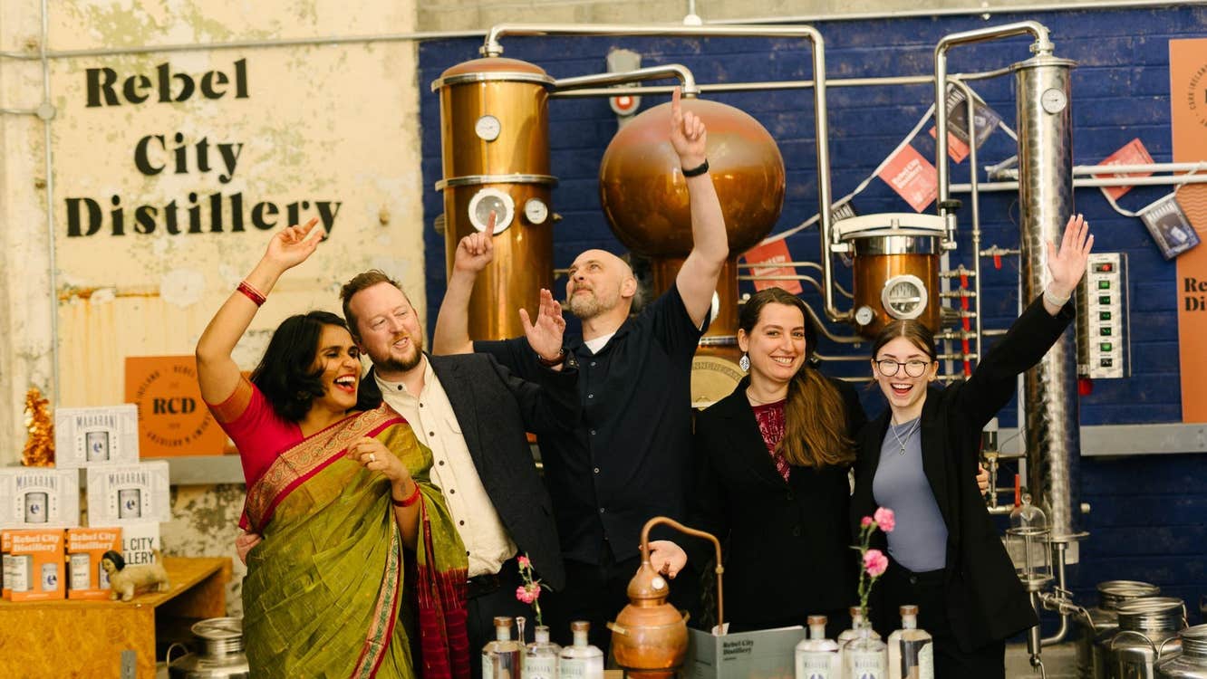 Five people with raised arms standing in a distillery and a counter with bottles on it in front