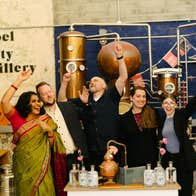 Five people with raised arms standing in a distillery and a counter with bottles on it in front