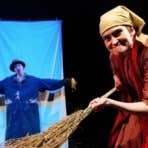 A woman acting as an old woman sweeping with a large twig broom.