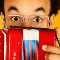 A man in glasses with raised eyebrows peering over the top of a small accordion he's holding up in front of him.