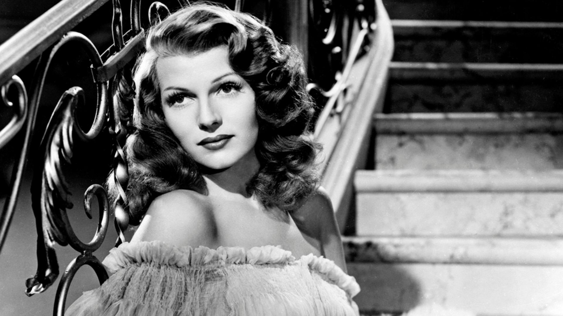 A glamorous woman is standing in front of a staircase.