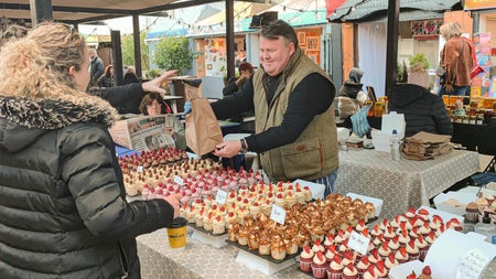 Person behind a stall with trays of little cakes handing a brown bag over to a lady
