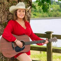 Effie Neill Dance - Gleneagle Arena, a smiling woman playing a guitar, leaning against a tree.
