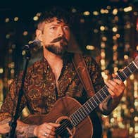 Niall McCabe to play Gleneagle Arena- a man playing a guitar standing at a mic on on stand with dark and gold blurry background.