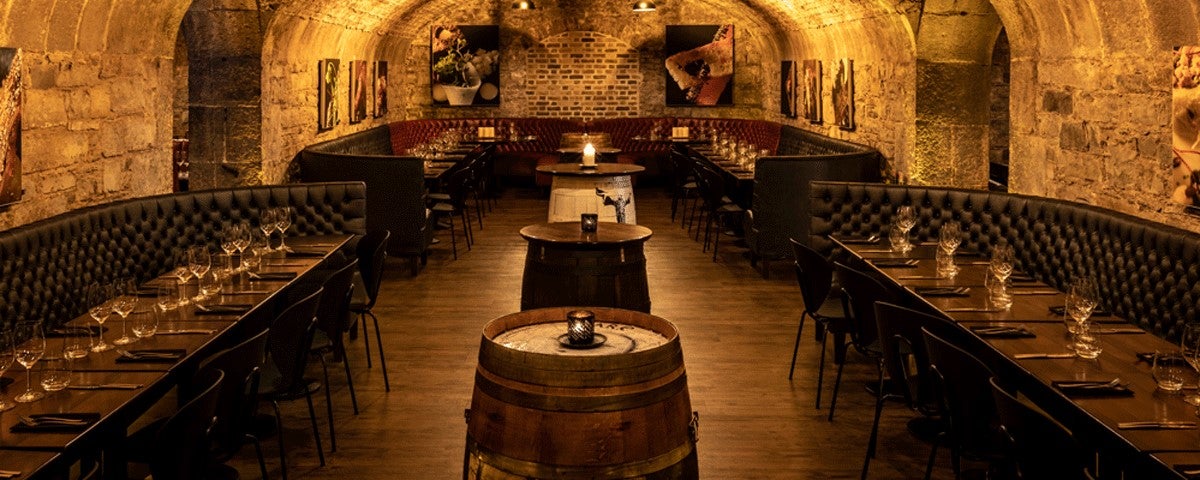 A stone walled room with a vaulted ceiling with wooden tables and beer barrels 