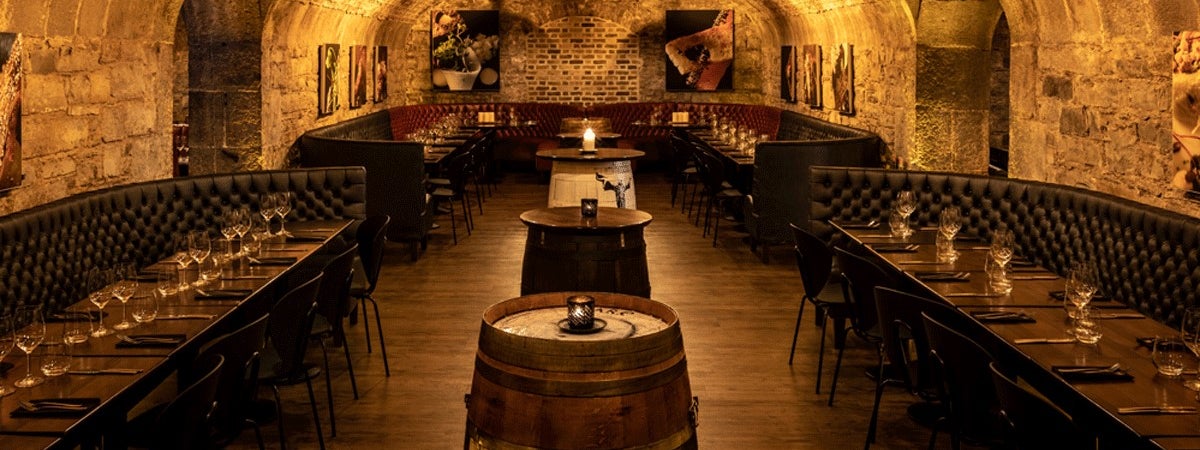 A stone walled room with a vaulted ceiling with wooden tables and beer barrels