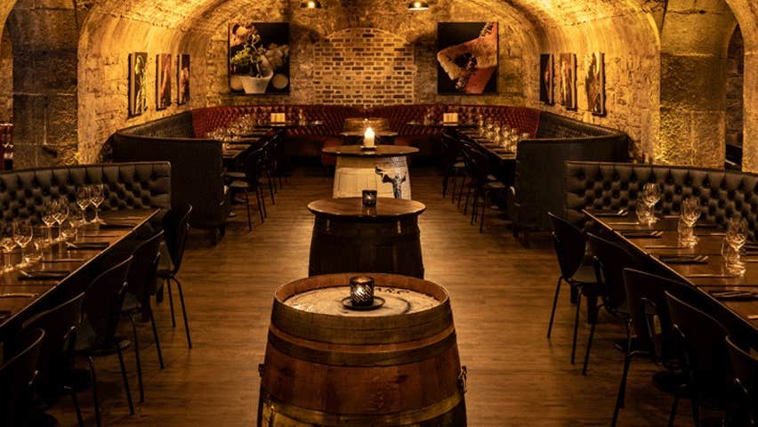 A stone walled room with a vaulted ceiling with wooden tables and beer barrels