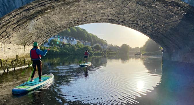 Two ladies stand up paddle boarding under a bridge with Cork City SUP