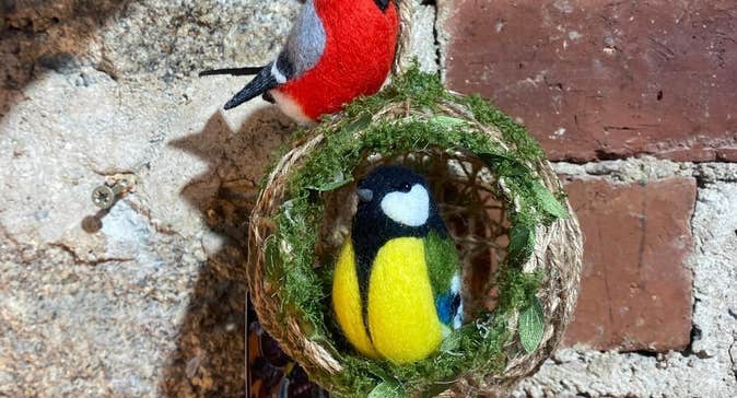 A felt brightly coloured bird in a small round woven half open ball with another felt brightly coloured bird on the top.