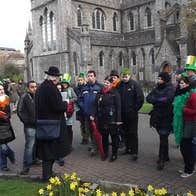 A man in dark overcoat and hat talking to a group of people outside a large, grey stone church.