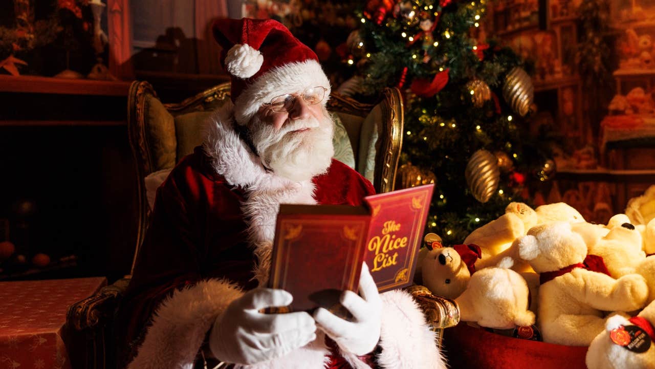 A santa dressed in dark red and white is reading a book with pile of stuffed toys beside him.