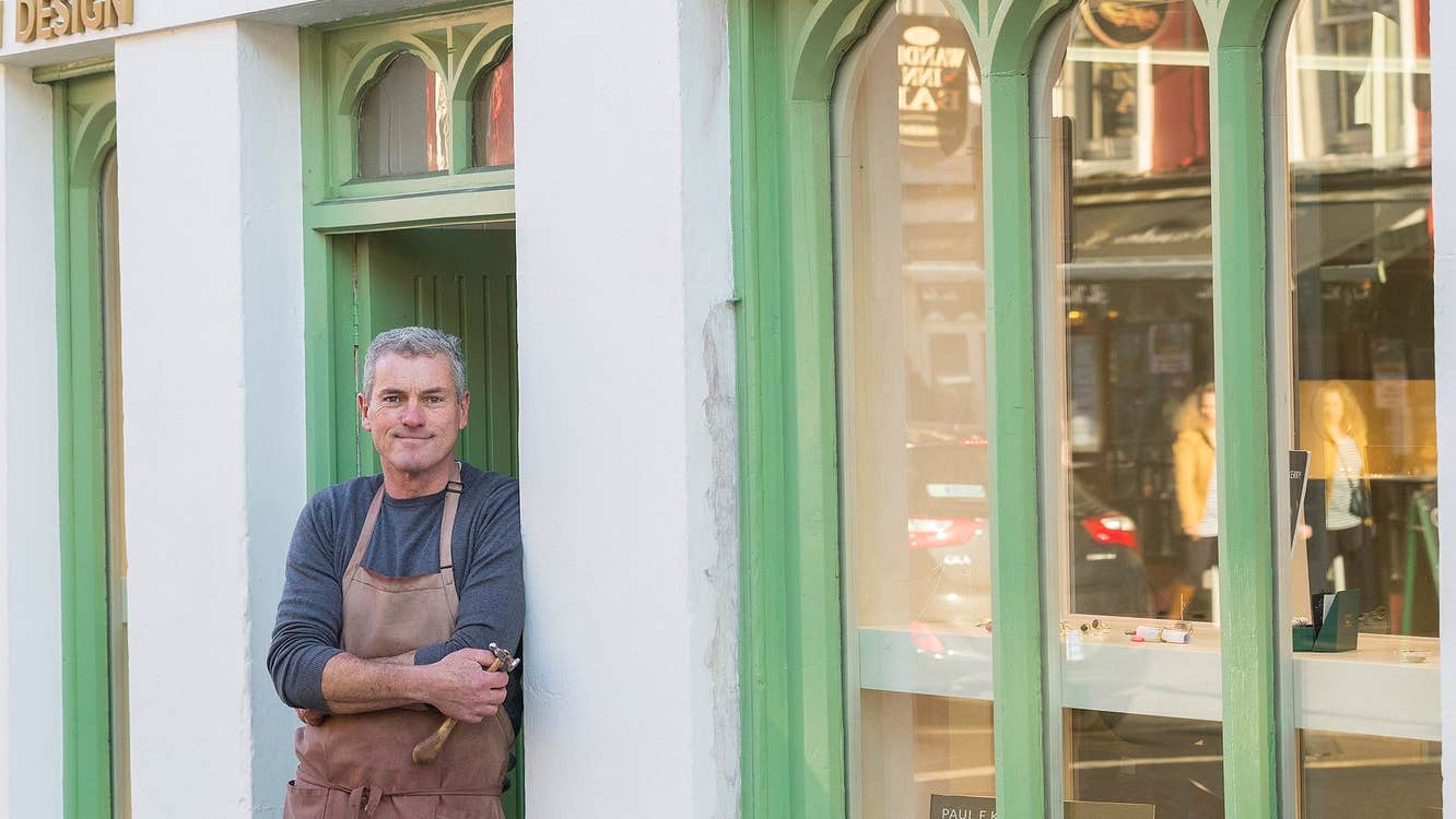 Man in a navy tee shirt and apron leaning against a shop doorway with windows to the right and pfk gold and silversmith written above