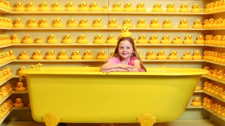 Rows of plastic ducks surrounding a yellow bath with a child in it and a duck on her head