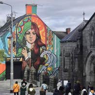 Cruinniú na nÓg 2026 Walking With Legends Live, a group of people on a pavement looking up at the end of building covered in a mural of a woman.
