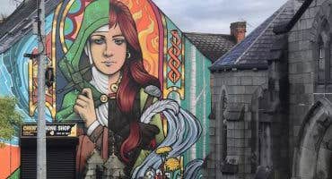 Cruinniú na nÓg 2026 Walking With Legends Live, a group of people on a pavement looking up at the end of building covered in a mural of a woman.