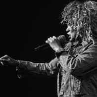 Side view of woman with big hair singing into a mic, against dark, plain background.