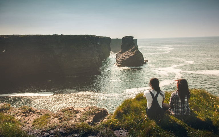 Two ladies sit on a grass verge on the coast looking across at cliffs in the distance