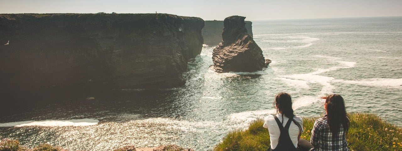 Two ladies sit on a grass verge on the coast looking across at cliffs in the distance