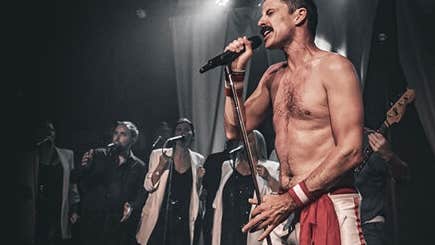 A man with no shirt on is singing with passion into a mic with singers in the background.