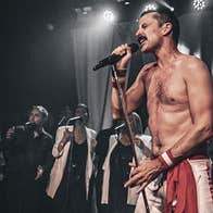 A man with no shirt on is singing with passion into a mic with singers in the background.