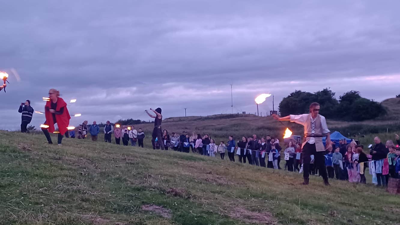 A large group of people in the distance on a hill at dusk, some holding lit flames.