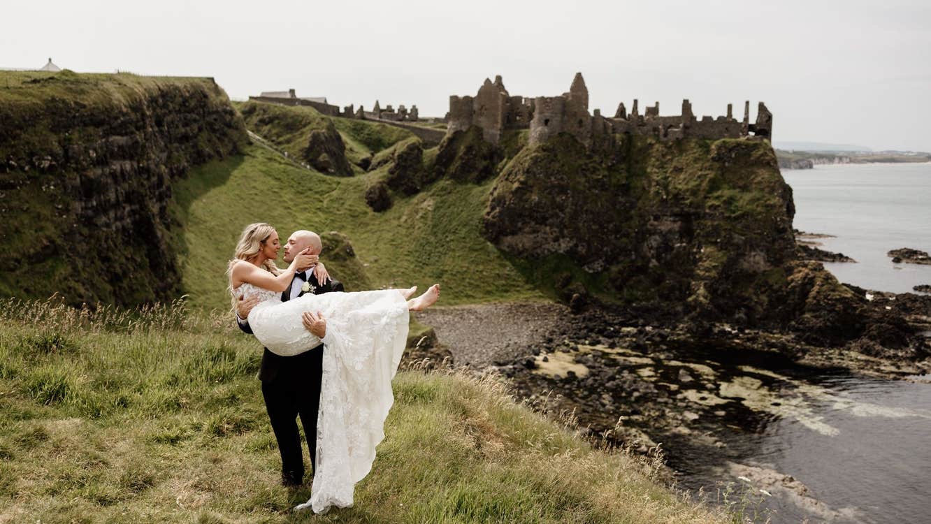 Groom holds bride in his arms with castle ruins and the sea in the background