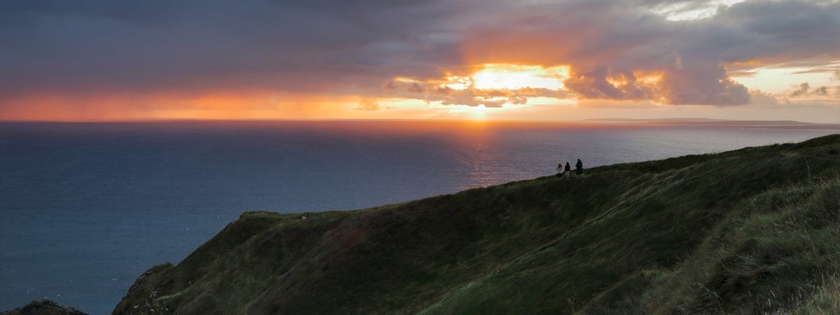 People stand on a cliff area looking out at a sunset