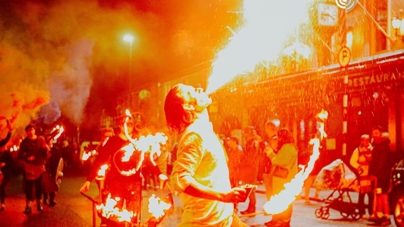 Orange coloured photo of person breathing fire with other lit objects in the background and people looking on.