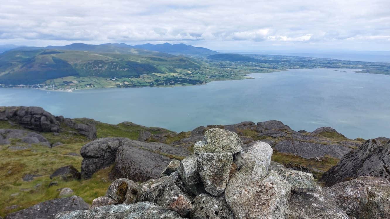 View from above a body of water taken from behind big stones with green fields in the distance