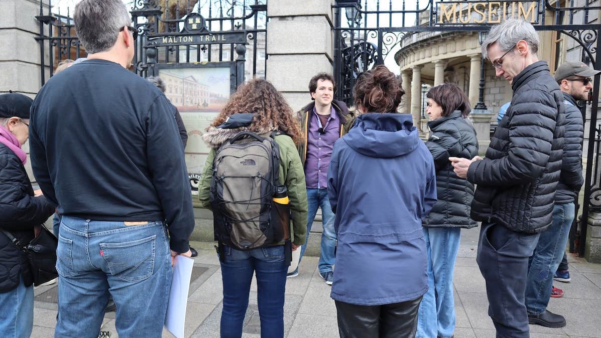A small group of people are standing listening to a man standing outside tall railings and cream stone wall.