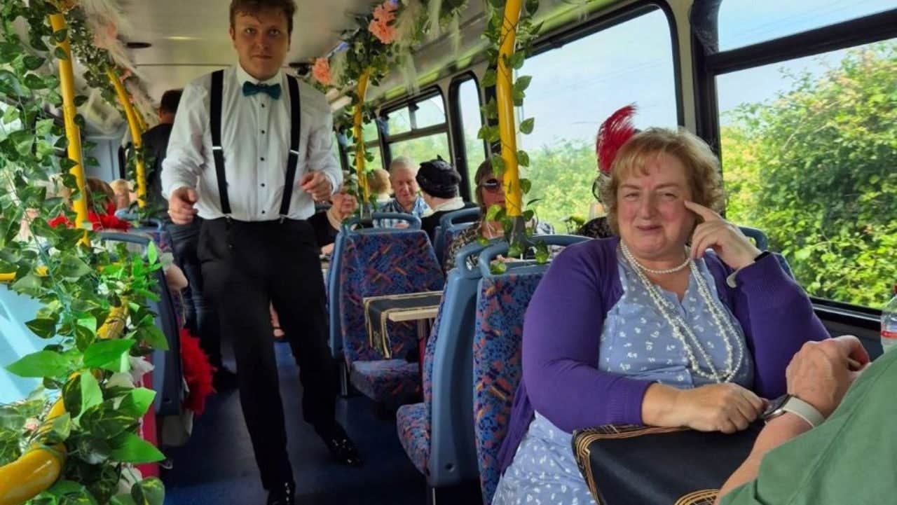 A man stands in the aisle of an Emerald Tea Tours bus with a woman smiling and sitting at a table by the window