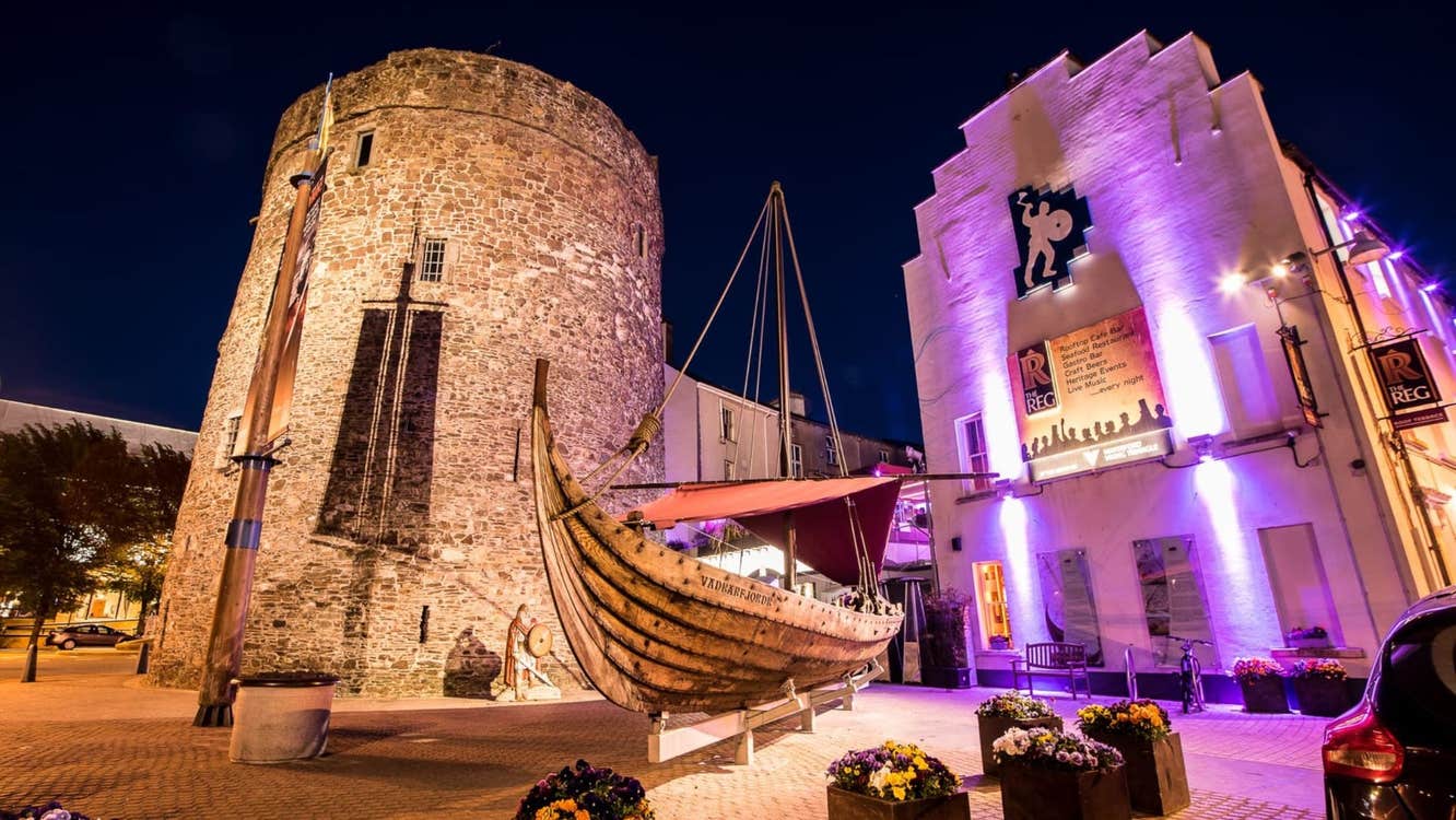 Outside The Reg Waterford lit up at night with pink lights next to Reginald's Tower and a wooden ship