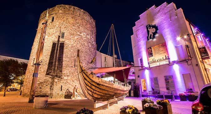 Outside The Reg Waterford lit up at night with pink lights next to Reginald's Tower and a wooden ship