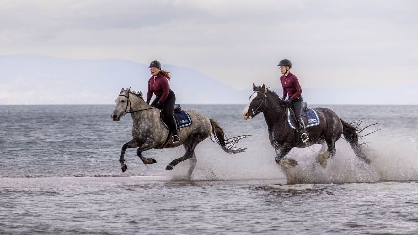 Two people wearing helmets horse riding through water