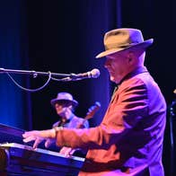 My Leonard Cohen with Stewart D’Arrietta - a side view of a man in a hat with a brim playing a piano on a stage with other musicians.