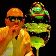 A man in dark glasses is blowing through a small tube with multiple large bubbles at the end