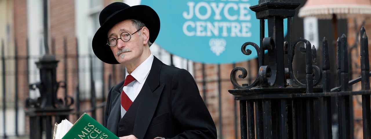 An elderly man in black suit and hat is holding open a green book, standing in front of black railings.