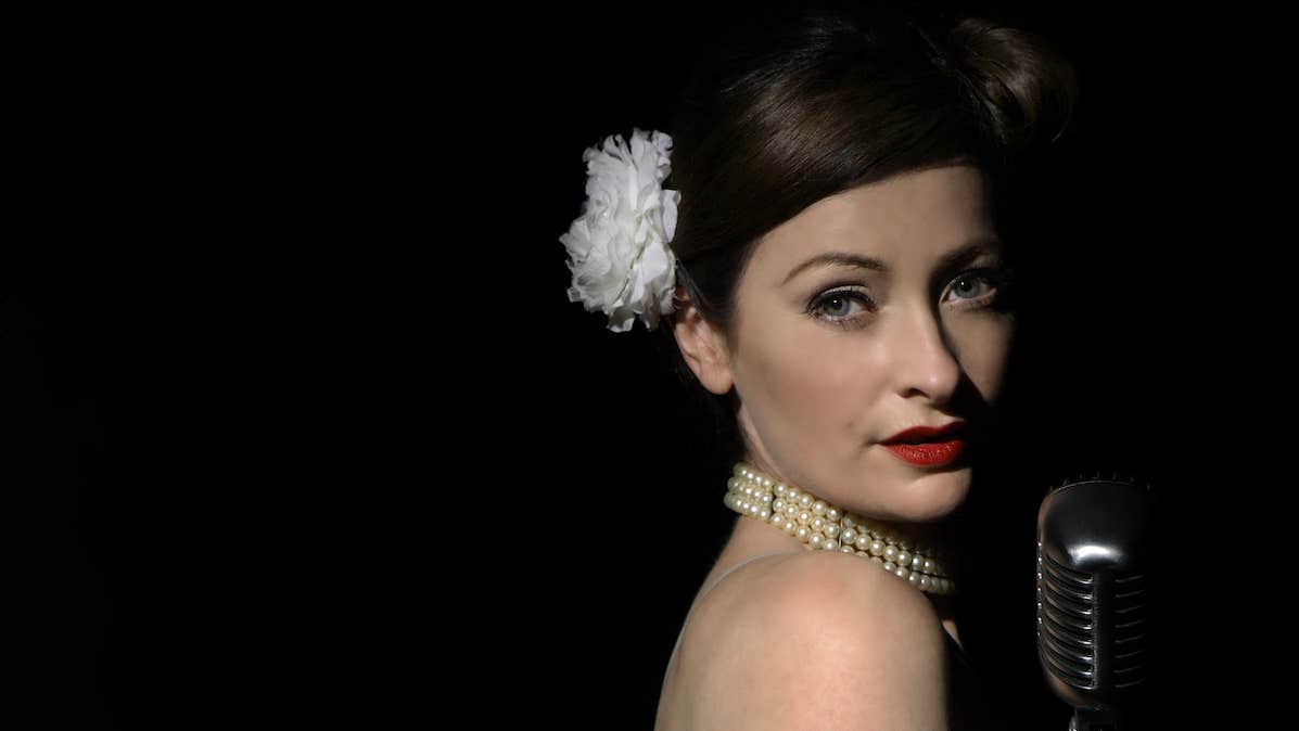 A woman with tied back hair held by white flower is looking over her bare right shoulder at camera with an old style mic in front of her, against black background.