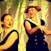 2 women on a stage, on seated at a piano singing, the other dancing beside with gold background.